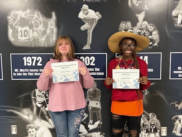 EAJ Student of the Week Emma Coburn and Allizah Moore holding their certificate in front of the Mt. Morris Athletic mural.