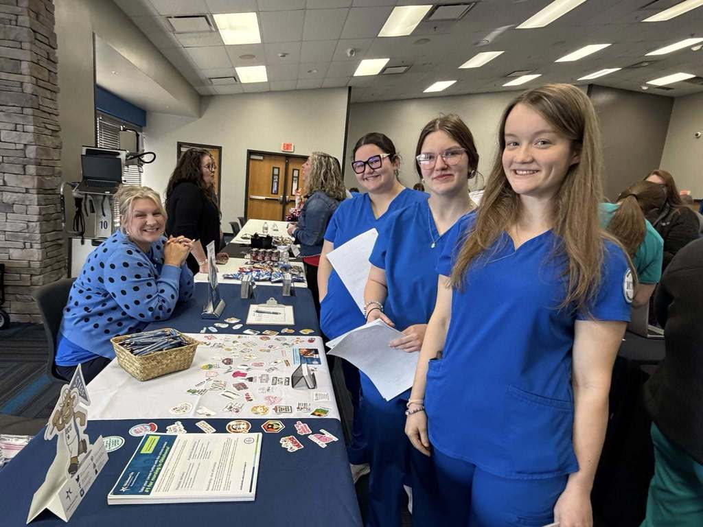 Four women in blue scrubs stand behind a table with items, papers, and a basket.