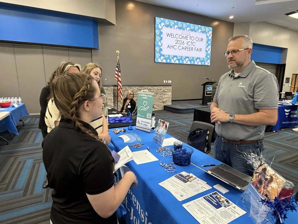 A person in a gray shirt speaks to a woman in a black shirt at a blue table with pamphlets.