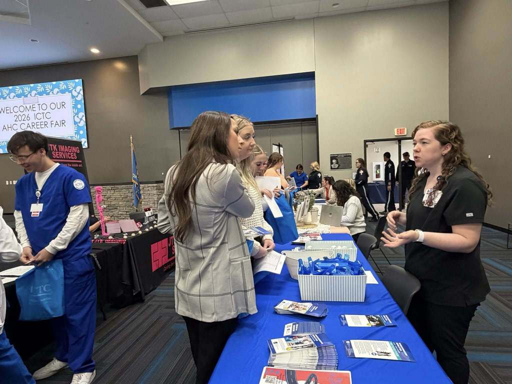 A group of people in white coats and blue scrubs are at a table with items. A screen is visible in the background.