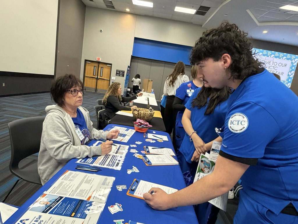 Four people in matching blue shirts sit at a table with printed materials. One person holds a pen.