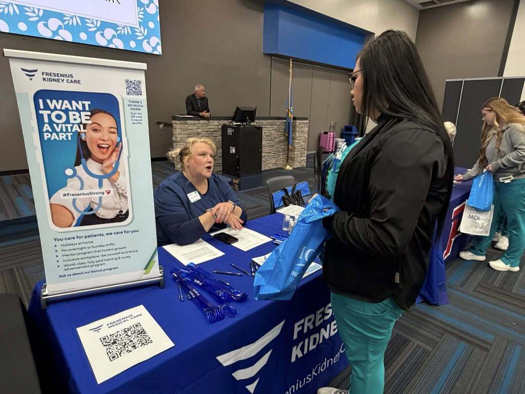 Two women interact at a table with a sign that says "I Want to Be a Vital Part". A large banner is behind them.