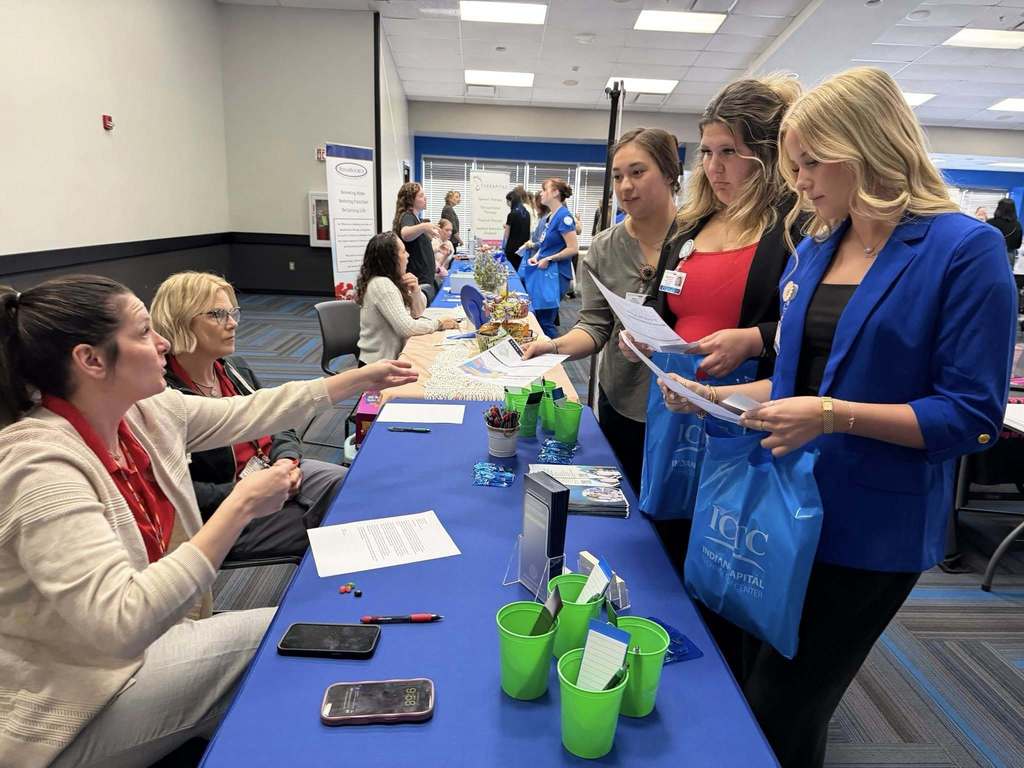 People stand at a table with blue tablecloths and green cups. A woman speaks with others while holding papers.