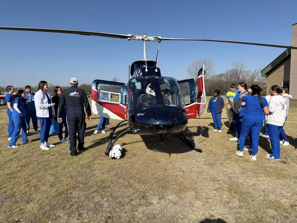Several people in blue uniforms surround a black helicopter with red and white accents on a grassy field.