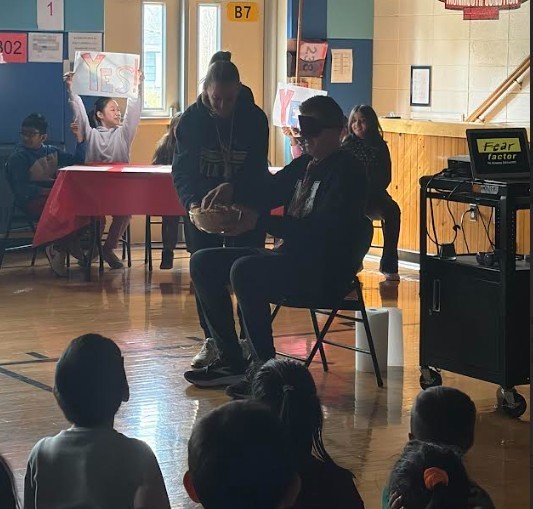A classroom with students sitting and standing. A man is seated on a chair with a baseball glove.