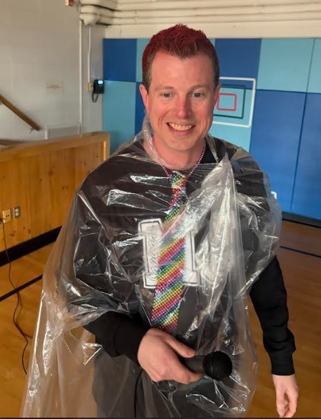 Man with red hair in plastic poncho and tie holding microphone, smiling. Behind him, a blue wall and wooden platform.
