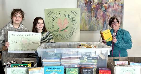 An adult and two students holding books, and a sign that says Cozy Corner's Book Drive and there is a lot of books in front of all of them. 