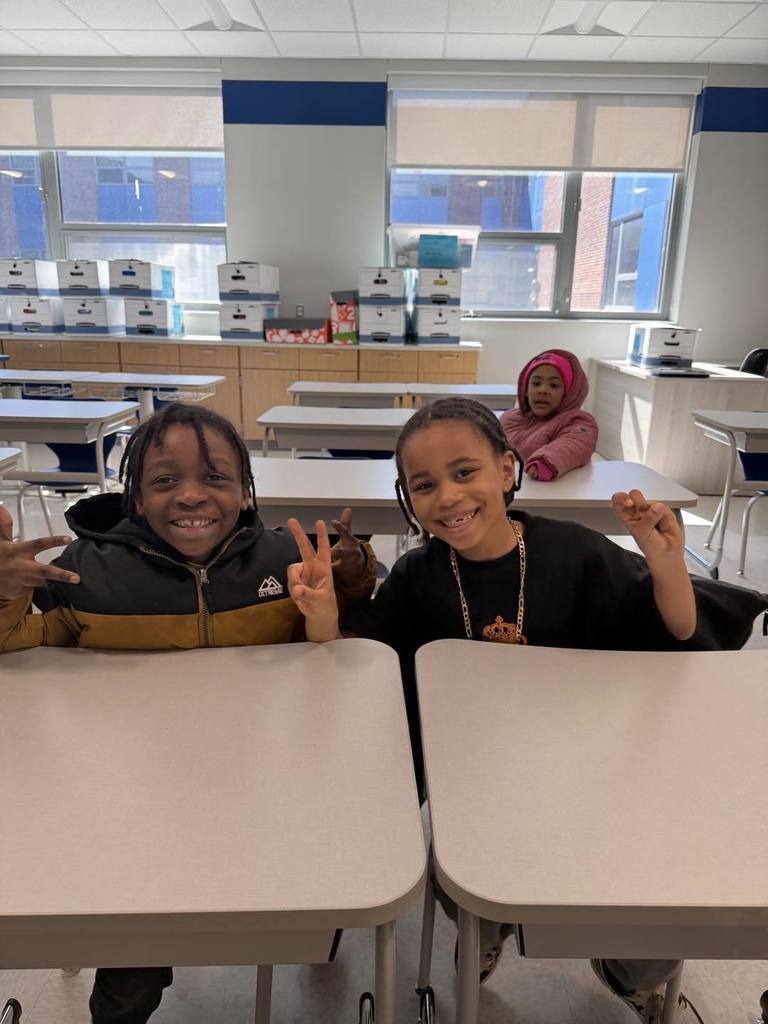 Two young boys sitting in their new classroom and smiling while they hold up peace signs. 