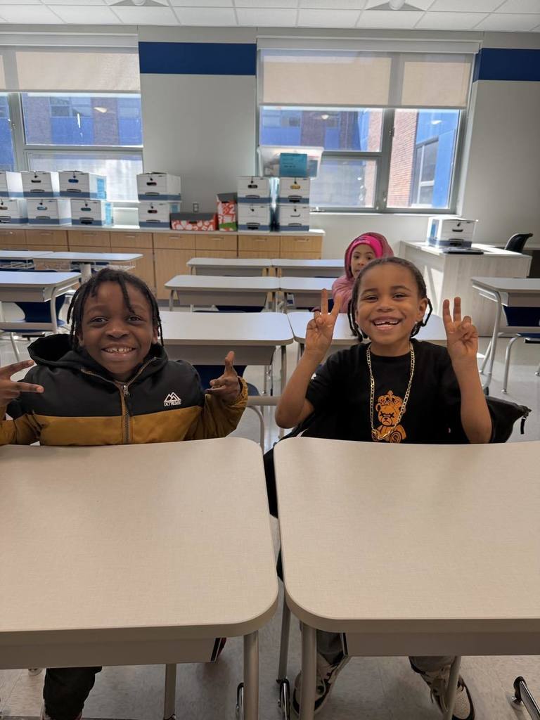 Two young boys sitting in their new classroom and smiling while they hold up peace signs. 