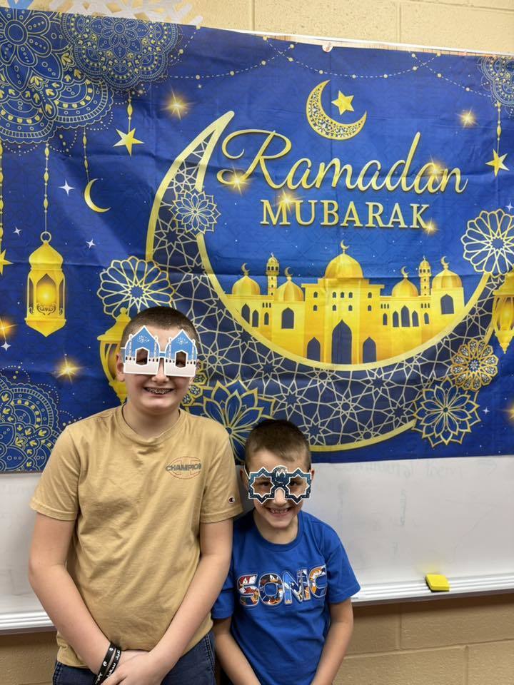 Students wearing festive glasses and standing in front of a Ramadan Mubarek banner. 