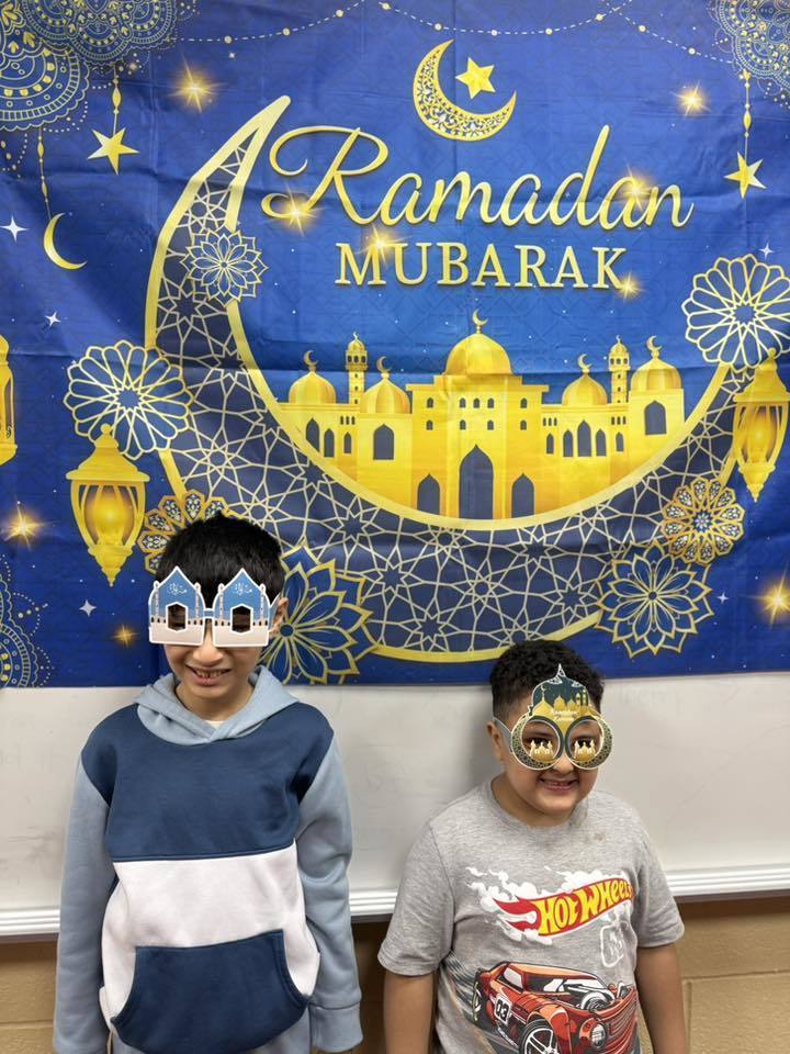 Students wearing festive glasses and standing in front of a Ramadan Mubarek banner. 