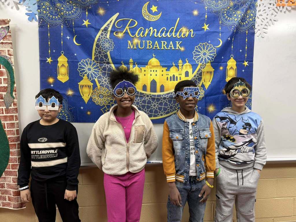 Students wearing festive glasses and standing in front of a Ramadan Mubarek banner. 