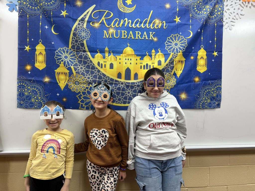 Students wearing festive glasses and standing in front of a Ramadan Mubarek banner. 