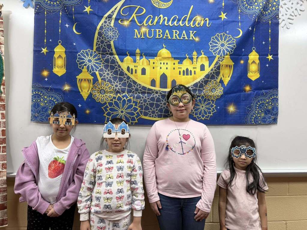 Students wearing festive glasses and standing in front of a Ramadan Mubarek banner. 