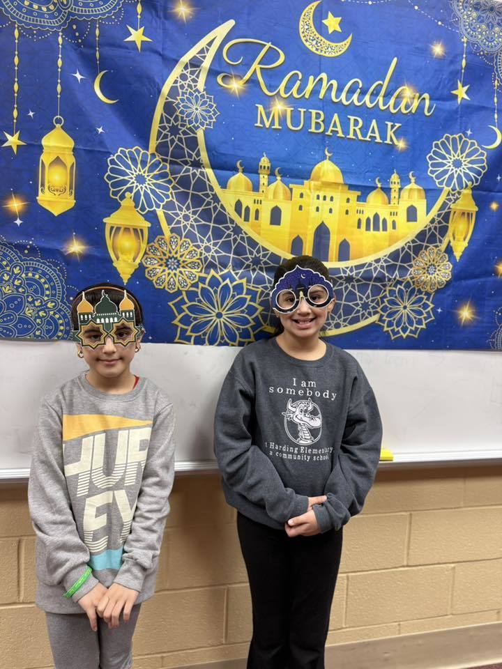 Students wearing festive glasses and standing in front of a Ramadan Mubarek banner. 