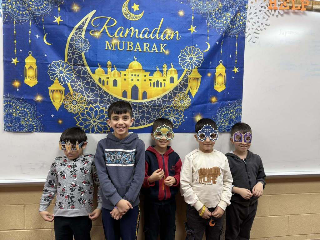 Students wearing festive glasses and standing in front of a Ramadan Mubarek banner. 