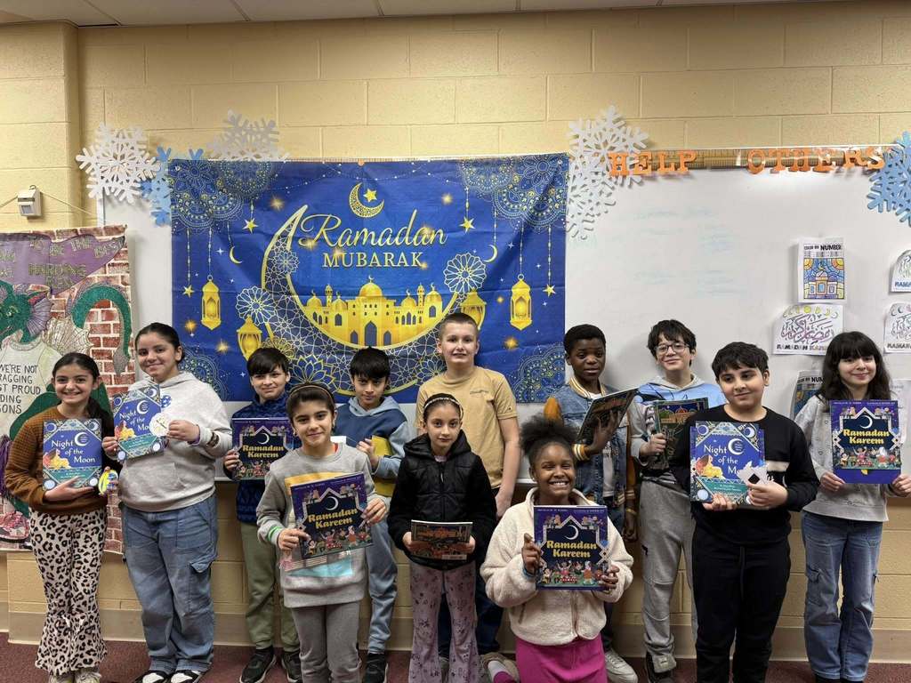 Students holding books for Ramadan and standing in front of a Ramadan Mubarek banner. 