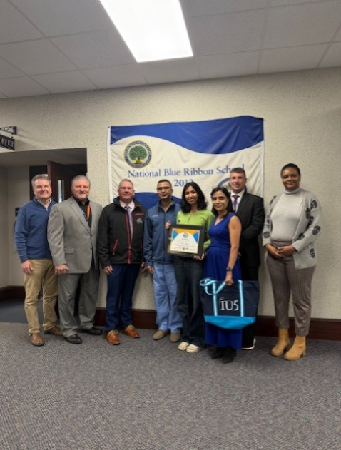 Young girl holding her award is standing next to her parents, sponsors, and news cast. 
