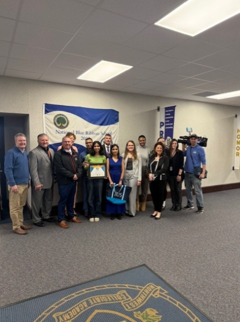 Young girl holding her award is standing next to her parents, school staff, sponsors, and news cast. 