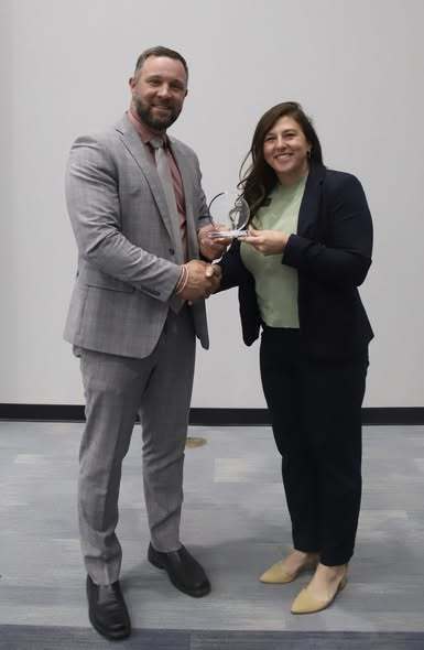 A man and a woman stand in a room, holding a small glass award, smiling and looking at the camera.
