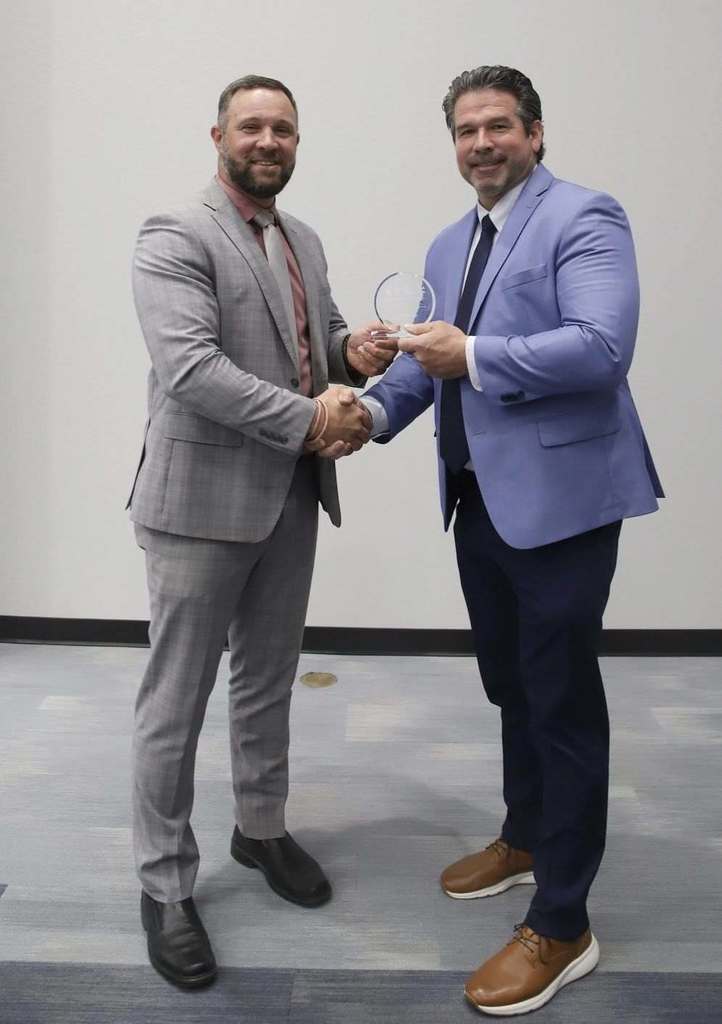 Two men in suits and ties, one in gray and the other in blue, shake hands and hold a trophy.
