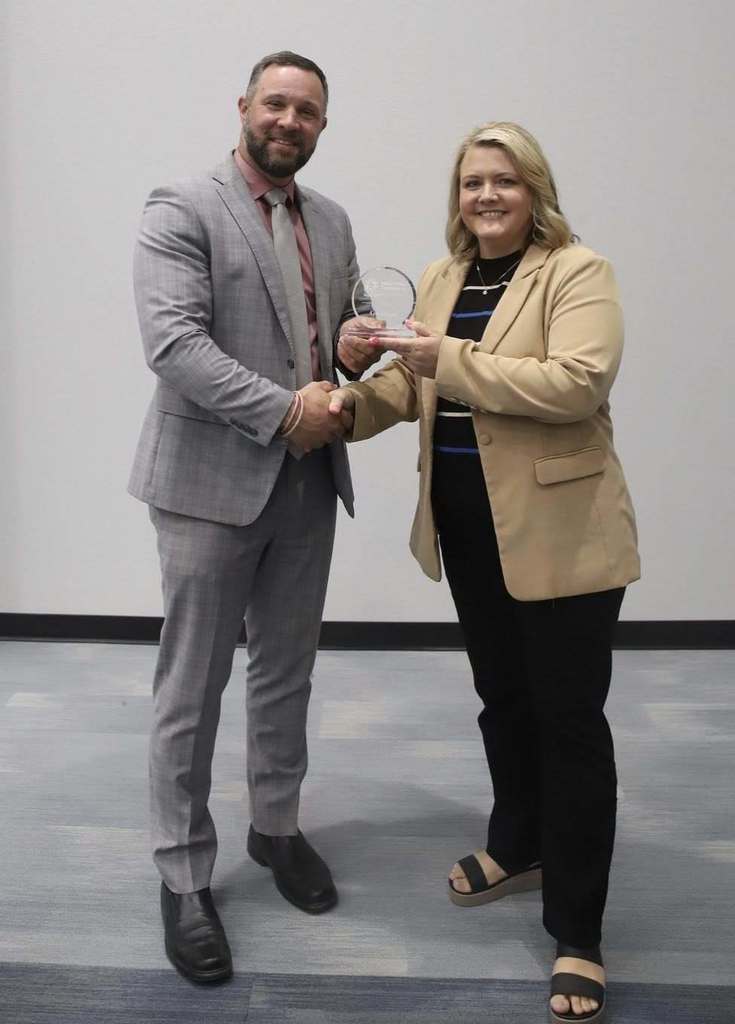 A man and woman exchange a glass trophy between them in a meeting room.