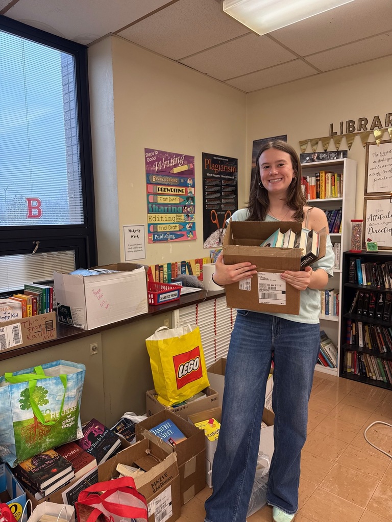 Aly holding a box of books with hundreds more in boxes on the floor