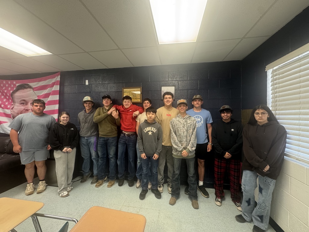 Nine individuals are standing in a room with a blue wall, American flag, and two tables. One person is sitting.