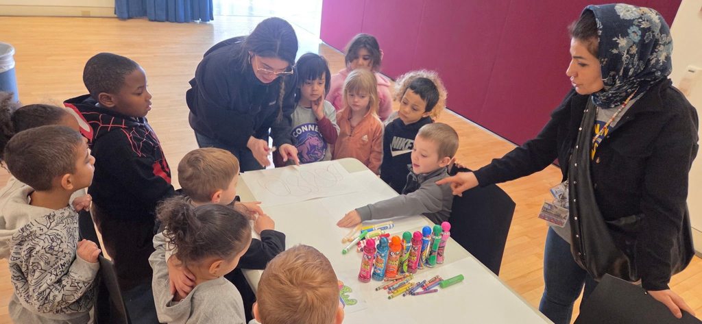 Kids gathered around the table watching a women draw art. 