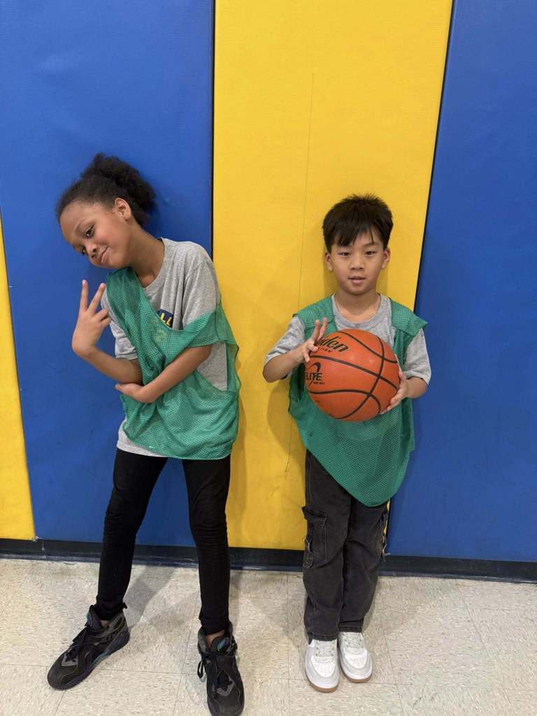 Two students wearing green jerseys over their clothes are both holding up two fingers. 