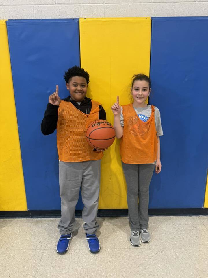 Two students wearing orange jerseys over their clothes are both holding up their pointer finger. 