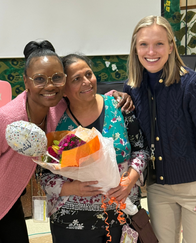 Three women stand together; the middle woman holds a bouquet of flowers, and the one on the right smiles.