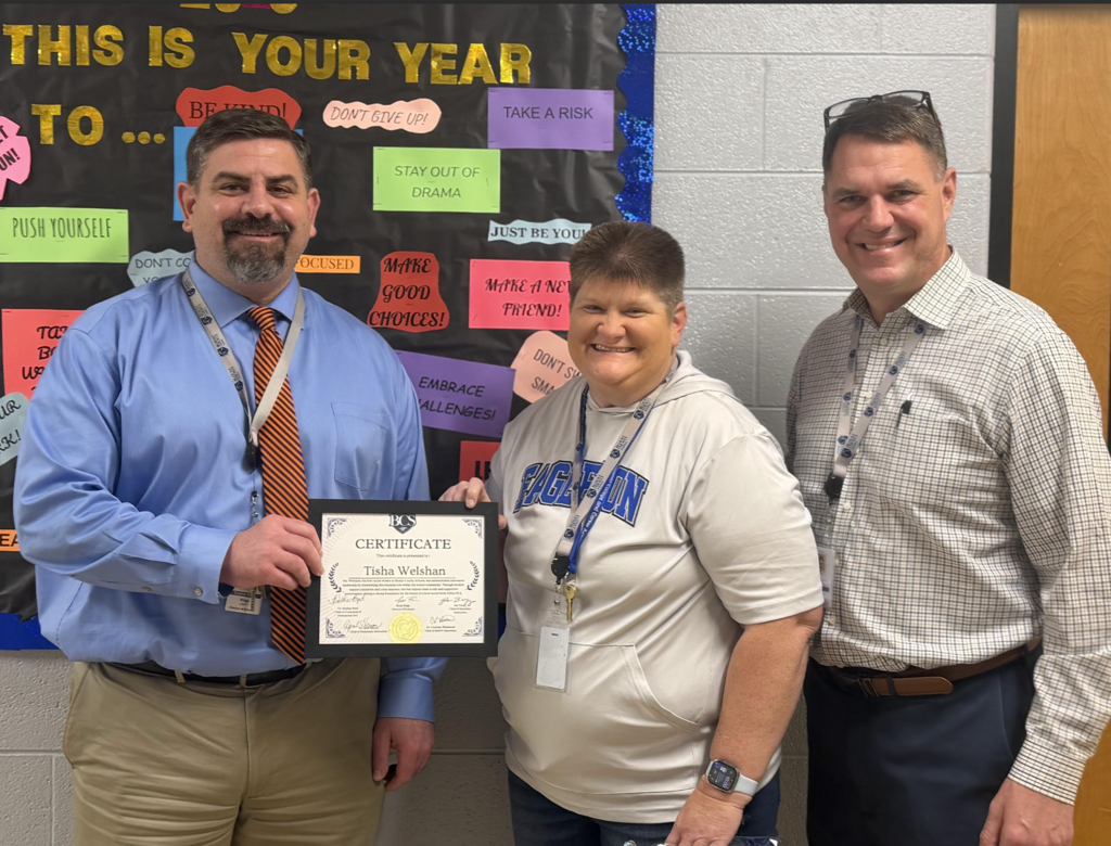 Image Description: Tisha Welshan, wearing a grey "Eagles" hoodie, smiles while holding her "Leader of Innovation" certificate. She is standing between two BCS administrators in front of a classroom bulletin board that says "This Is Your Year" and "Be Kind." The official Blount County Schools shield logo is placed in the corner of the image.