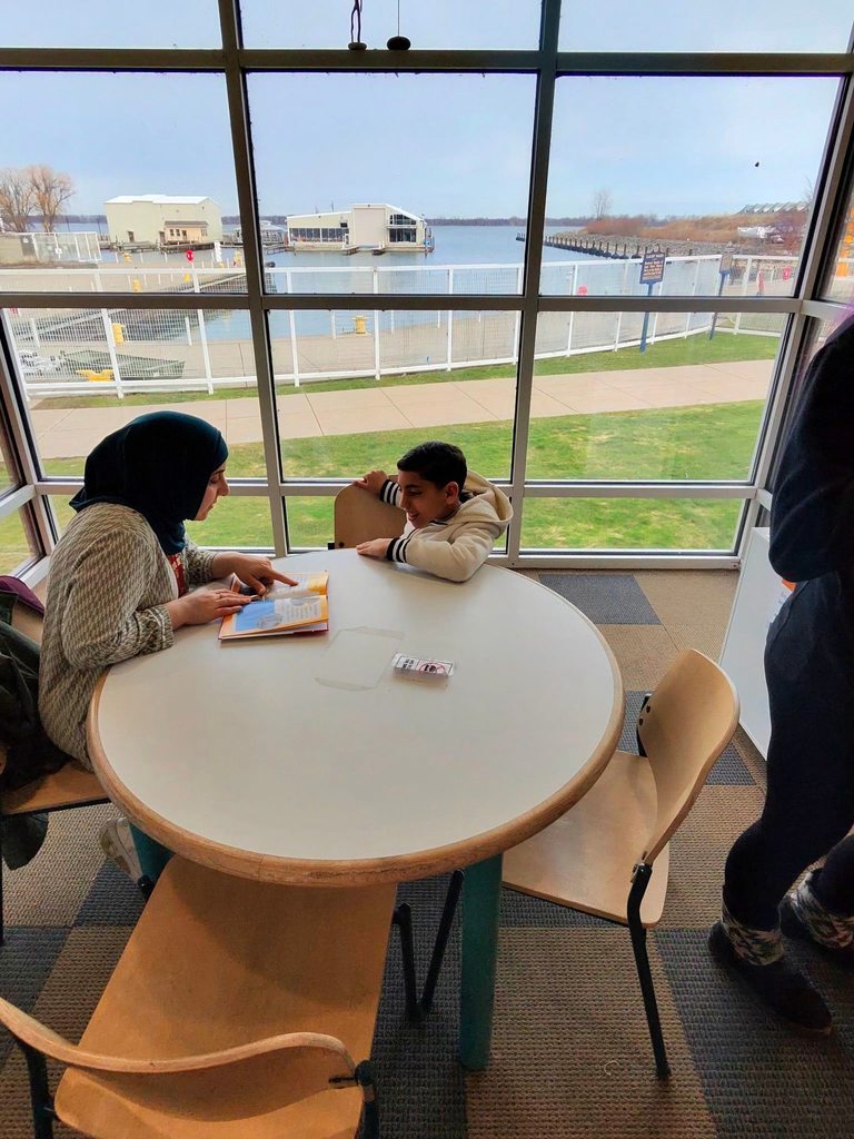 A women reading a book to a student in a while jacket in front of a large window that overlooks the water in the children's library. 