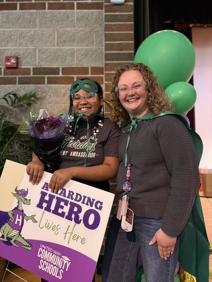 A mom is standing next to their child who has a green is holding a sign that says "A Harding Hero Lives Here" on it. 