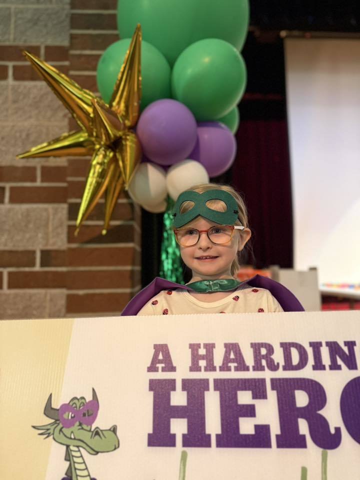 A child has a green is holding a sign that says "A Harding Hero Lives Here" on it. 