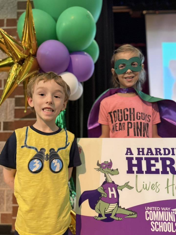 A young child with a yellow shirt on is standing next to a child who has a green is holding a sign that says "A Harding Hero Lives Here" on it. 