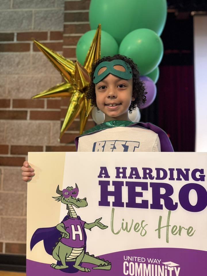 A child has a green is holding a sign that says "A Harding Hero Lives Here" on it. 