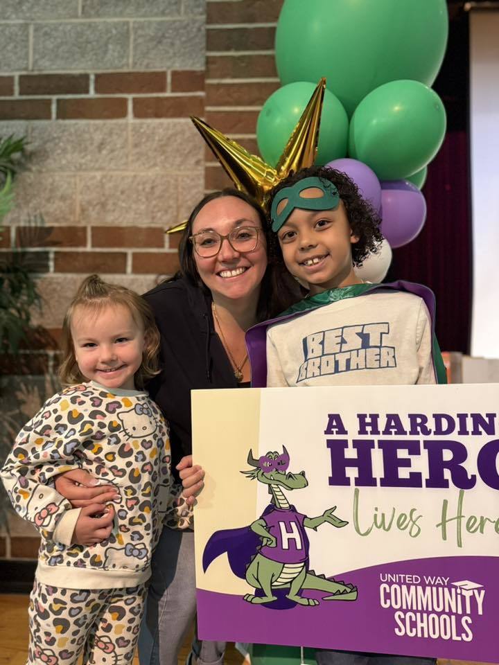 Mother and sister standing next to their child who has a green is holding a sign that says "A Harding Hero Lives Here" on it. 