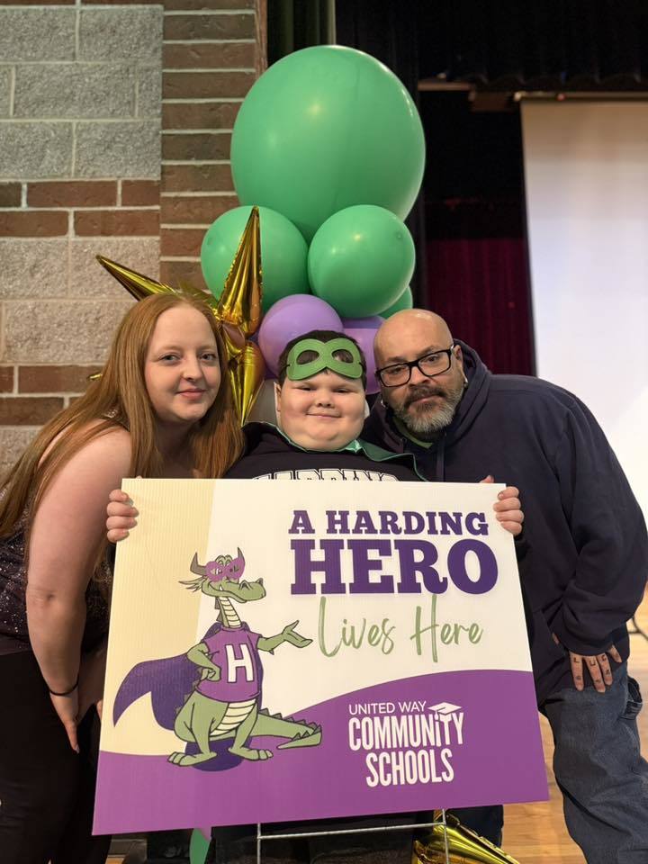 Parents standing next to their child who has a green is holding a sign that says "A Harding Hero Lives Here" on it. 