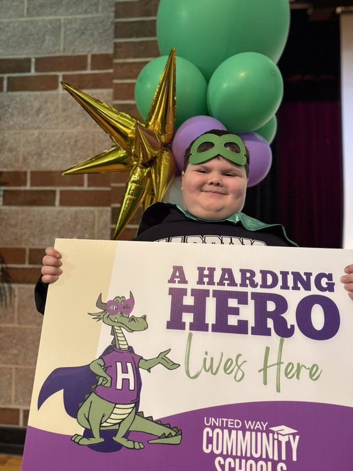 A child has a green is holding a sign that says "A Harding Hero Lives Here" on it. 