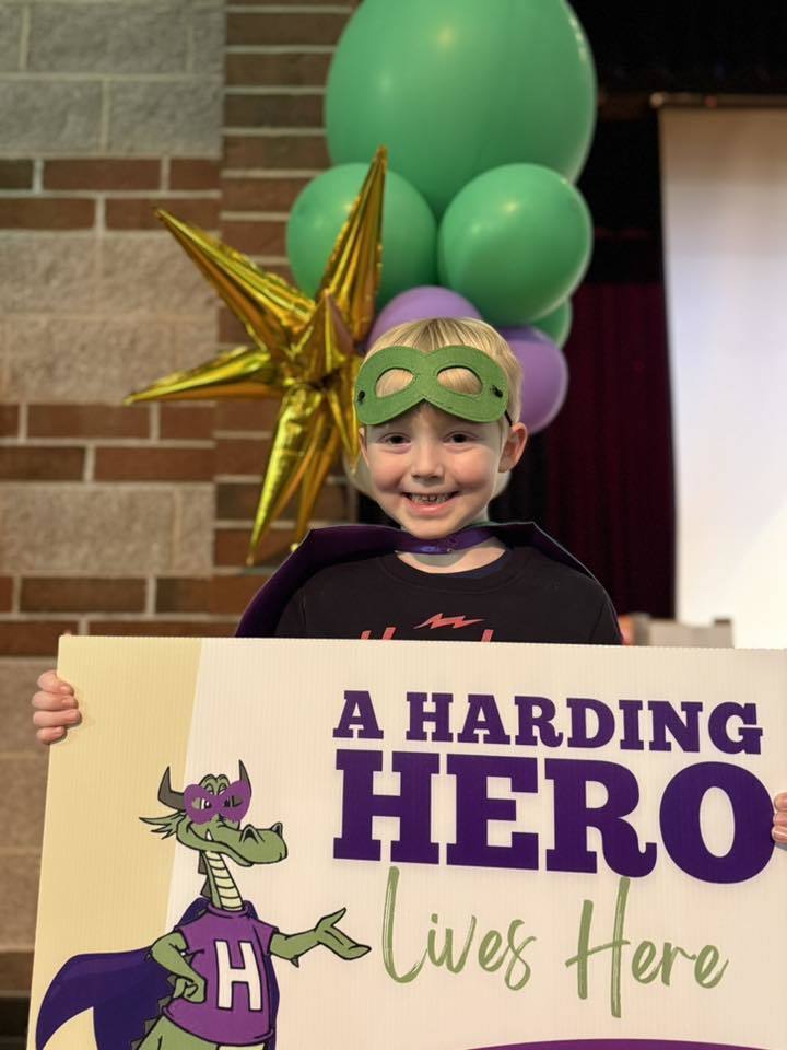 A child has a green is holding a sign that says "A Harding Hero Lives Here" on it. 