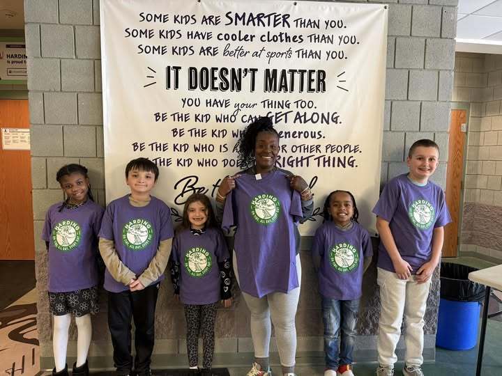 Students are wearing matching purple shirts with the Harding logo on them and a women in the middle is holding a matching shirt. 