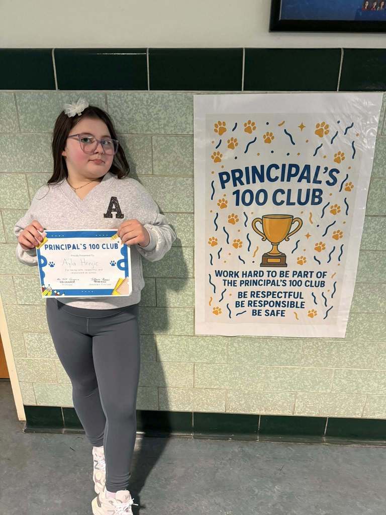 Young girl wearing a gray shirt with the letter "A" on it is holding his "Principal 100 Club" Certificate. 