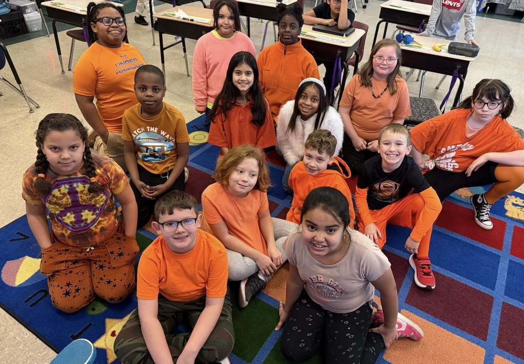 Students wearing orange and they are sitting on a colorful carpet in their classroom. 