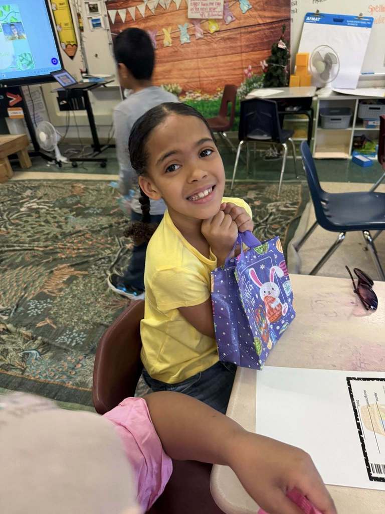 A young student is holding their Easter bag with goodies in it on their desk and smiling for a photo. 
