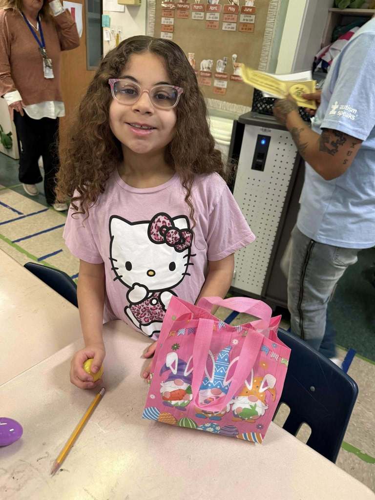 A young student is holding their Easter bag with goodies in it on their desk and smiling for a photo. 