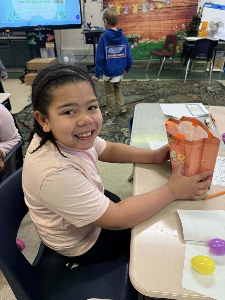 A young student is holding their Easter bag with goodies in it on their desk and smiling for a photo. 