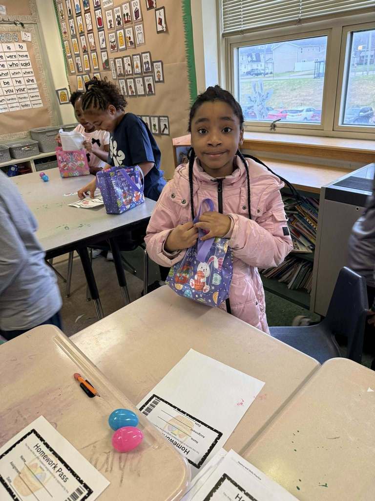 A young student is holding their Easter bag with goodies in it on their desk and smiling for a photo. 