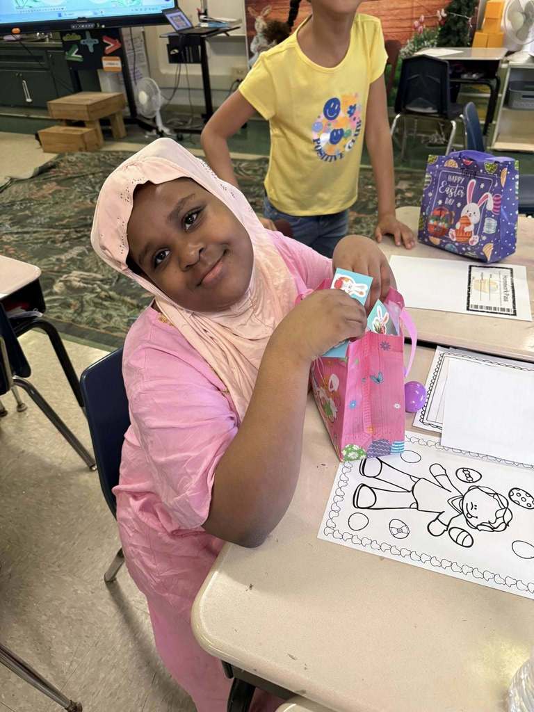 A young student is holding their Easter bag with goodies in it on their desk and smiling for a photo. 